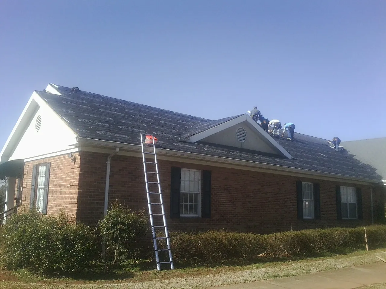 Crew tearing shingles off a brick ranch roof with a ladder set — Emergency Roof Repair work by our crew in Pine Hill